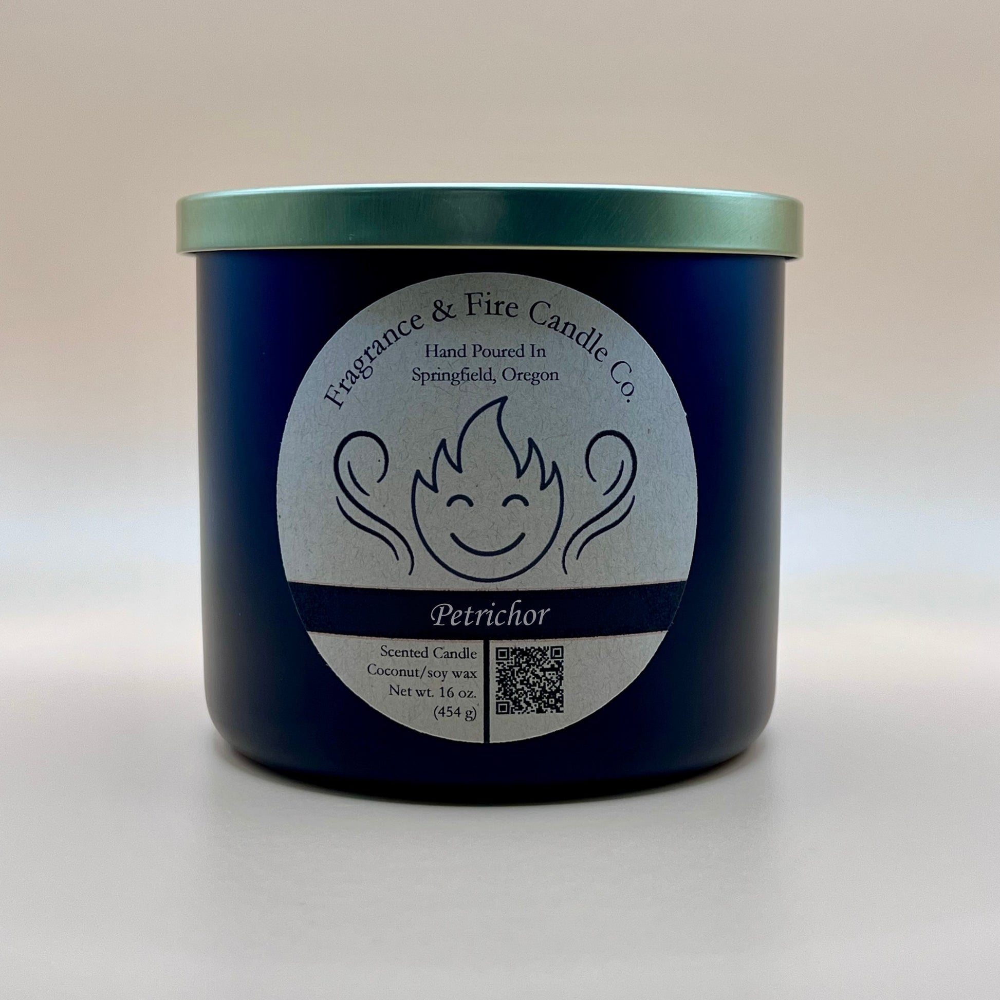 Black glass container candle from Fragrance & Fire Candle Co. with a gold lid. The label features a stylized flame with a smiling face, the text "Fragrance & Fire Candle Co. Hand Poured In Springfield, Oregon," and "Petrichor" scent name. Details include "Scented Candle Coconut/soy wax Net wt. 16 oz (454 g)" and a QR code.