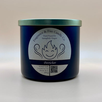 Black glass container candle from Fragrance & Fire Candle Co. with a gold lid. The label features a stylized flame with a smiling face, the text "Fragrance & Fire Candle Co. Hand Poured In Springfield, Oregon," and "Petrichor" scent name. Details include "Scented Candle Coconut/soy wax Net wt. 16 oz (454 g)" and a QR code.