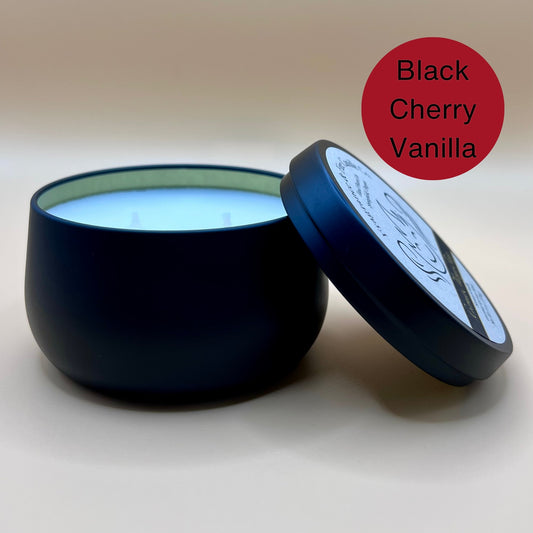 6oz black tin candle from Fragrance & Fire Candle Co., Springfield, Oregon, with an open lid resting beside it. The tin contains white wax. A dark red circle in the upper right corner reads "Black Cherry Vanilla." The open lid shows a partial label design.