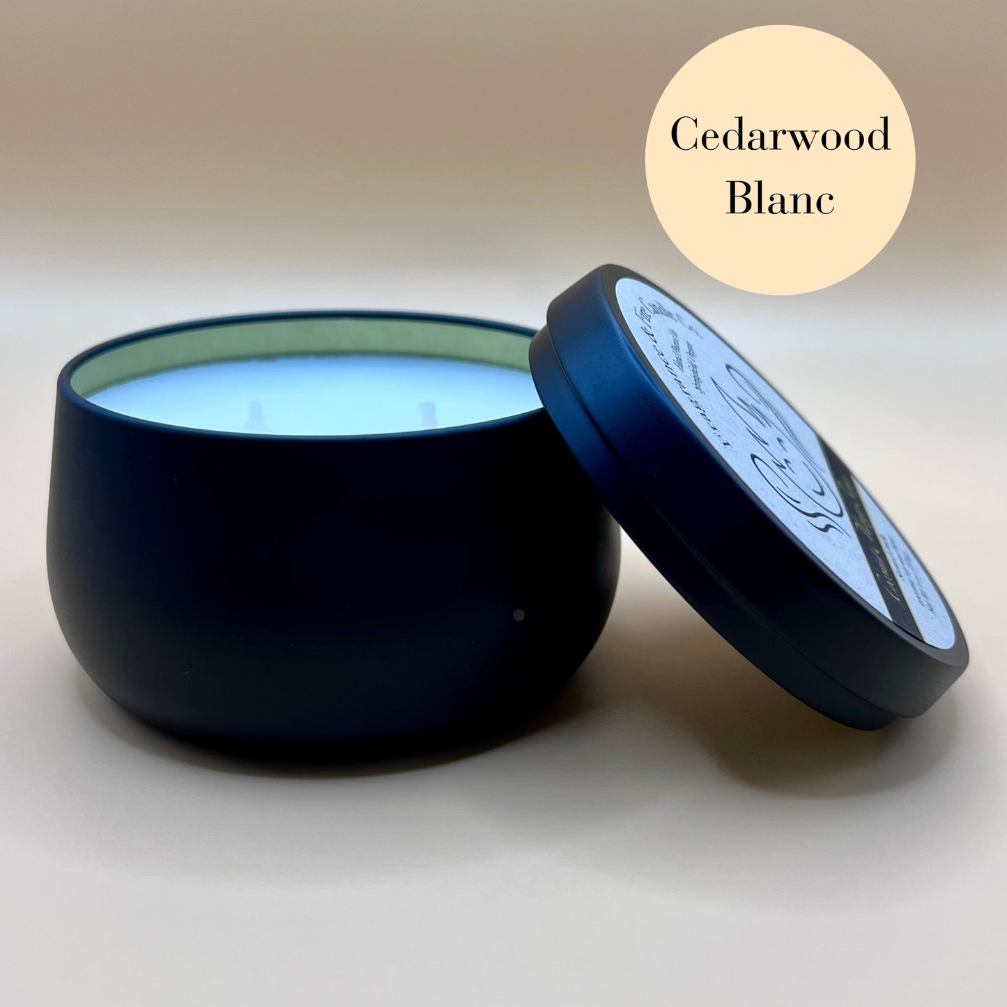 6oz black tin candle from Fragrance & Fire Candle Co., Springfield, Oregon, with an open lid resting beside it. The tin contains white wax. A cream circle in the upper right corner reads "Cedarwood Blanc." The open lid shows a partial label design.