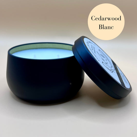 6oz black tin candle from Fragrance & Fire Candle Co., Springfield, Oregon, with an open lid resting beside it. The tin contains white wax. A cream circle in the upper right corner reads "Cedarwood Blanc." The open lid shows a partial label design.