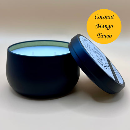 6oz black tin candle from Fragrance & Fire Candle Co., Springfield, Oregon, with an open lid resting beside it. The tin contains white wax. A yellow circle in the upper right corner reads "Coconut Mango Tango." The open lid shows a partial label design.