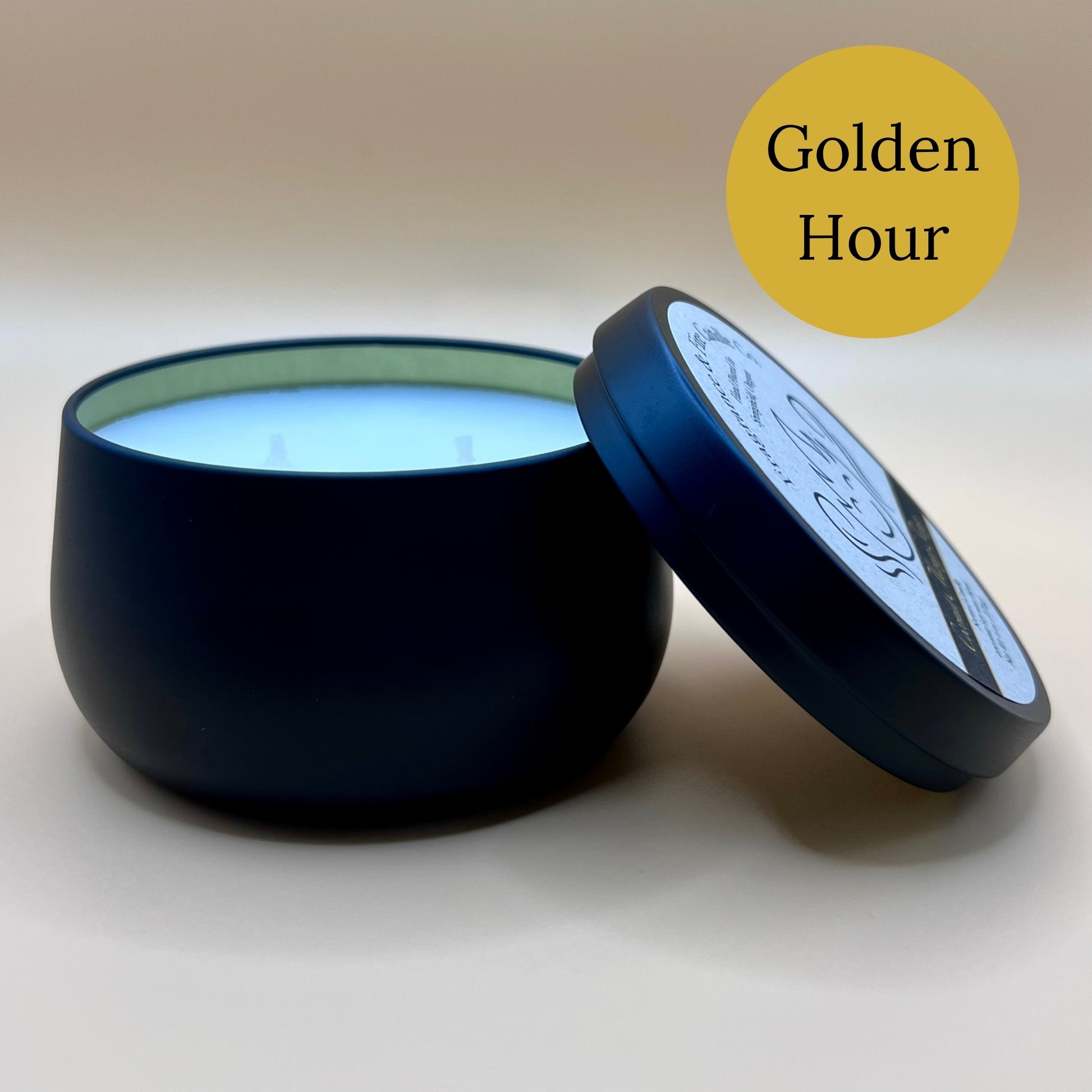 6oz black tin candle from Fragrance & Fire Candle Co., Springfield, Oregon, with an open lid resting beside it. The tin contains white wax. A dark yellow circle in the upper right corner reads "Golden Hour." The open lid shows a partial label design.