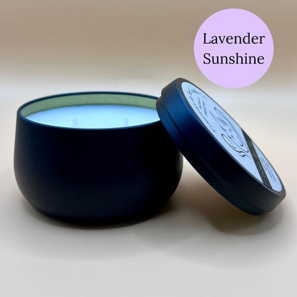 6oz black tin candle from Fragrance & Fire Candle Co., Springfield, Oregon, with an open lid resting beside it. The tin contains white wax. A light purple circle in the upper right corner reads "Lavender Sunshine." The open lid shows a partial label design.