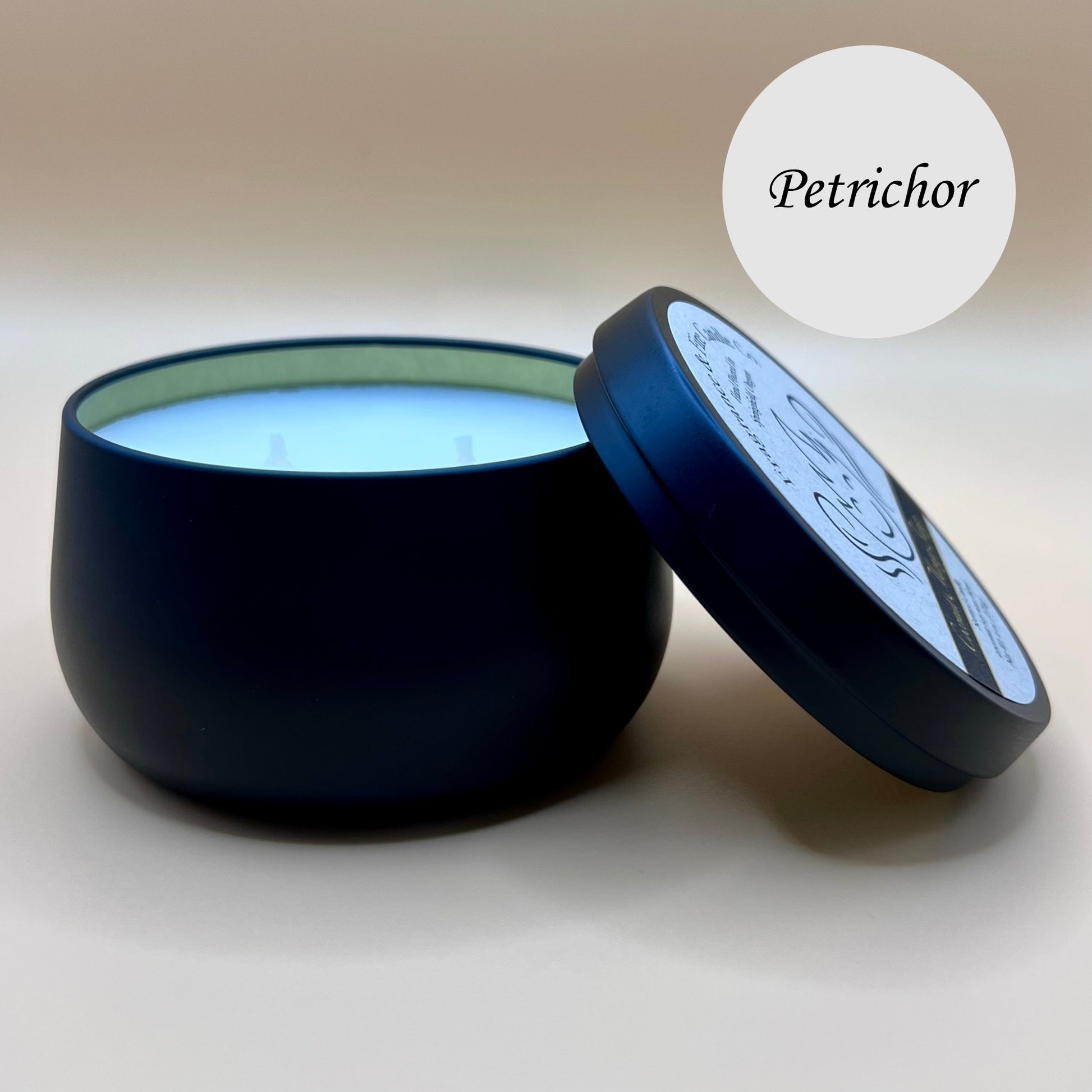 6oz black tin candle from Fragrance & Fire Candle Co., Springfield, Oregon, with an open lid resting beside it. The tin contains white wax. A light circle in the upper right corner reads "Petrichor." The open lid shows a partial label design.