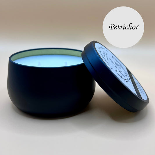 6oz black tin candle from Fragrance & Fire Candle Co., Springfield, Oregon, with an open lid resting beside it. The tin contains white wax. A light circle in the upper right corner reads "Petrichor." The open lid shows a partial label design.
