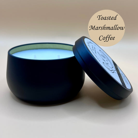 6oz black tin candle from Fragrance & Fire Candle Co., Springfield, Oregon, with an open lid resting beside it. The tin contains white wax. A tan circle in the upper right corner reads "Toasted Marshmallow Coffee." The open lid shows a partial label design.