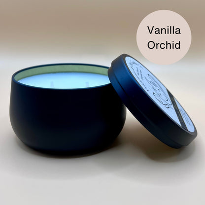 6oz black tin candle from Fragrance & Fire Candle Co., Springfield, Oregon, with an open lid resting beside it. The tin contains white wax. An off-white circle in the upper right corner reads "Vanilla Orchid." The open lid shows a partial label design.