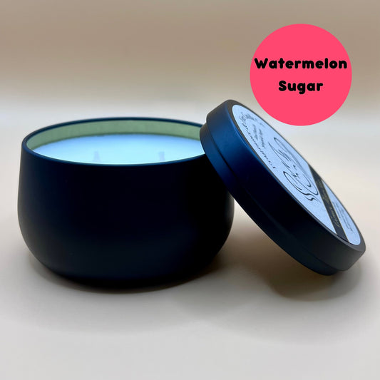 6oz black tin candle from Fragrance & Fire Candle Co., Springfield, Oregon, with an open lid resting beside it. The tin contains white wax. A pink circle in the upper right corner reads "Watermelon Sugar." The open lid shows a partial label design.