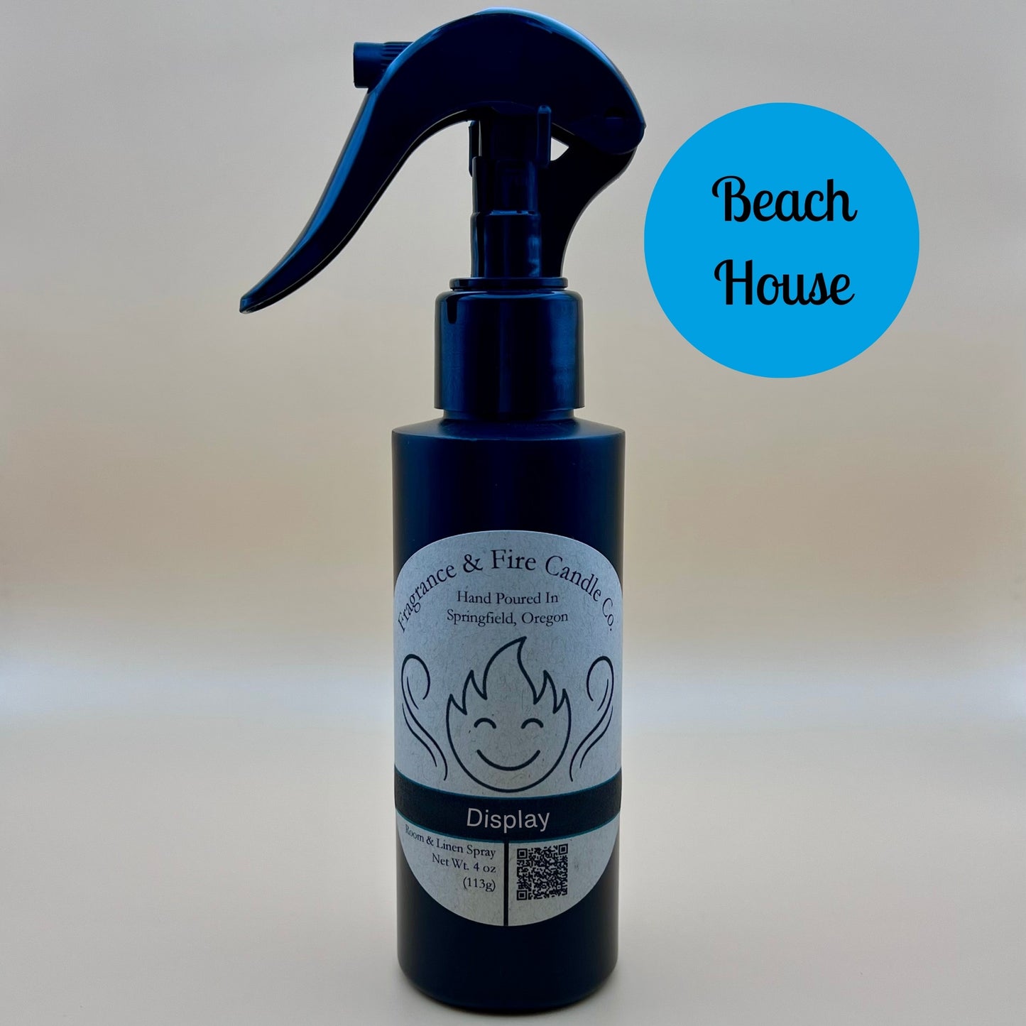 Black spray bottle from Fragrance & Fire Candle Co., Springfield, Oregon, with a black spray nozzle. The bottle has a white label with a stylized flame logo, the text "Fragrance & Fire Candle Co. Hand Poured In Springfield, Oregon," and "Display" scent name. The label also includes a QR code and the text "Room & Linen Spray Net Wt. 4 oz (113g)." A blue circle in the upper right corner reads "Beach House."