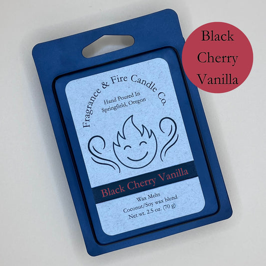 Black clamshell packaging for wax melts from Fragrance & Fire Candle Co., Springfield, Oregon. The packaging features a white label with a stylized flame logo, the text "Fragrance & Fire Candle Co. Hand Poured In Springfield, Oregon," and "Black Cherry Vanilla" scent name. The label also includes the text "Wax Melts Coconut/Soy wax blend Net wt. 2.5 oz. (70 g)." A red circle in the upper right corner says scent name.