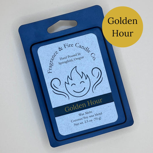 Black clamshell packaging for wax melts from Fragrance & Fire Candle Co., Springfield, Oregon. The packaging features a white label with a stylized flame logo, the text "Fragrance & Fire Candle Co. Hand Poured In Springfield, Oregon," and "Golden Hour" scent name. The label also includes the text "Wax Melts Coconut/Soy wax blend Net wt. 2.5 oz. (70 g)." A dark yellow circle in the upper right corner says scent name.