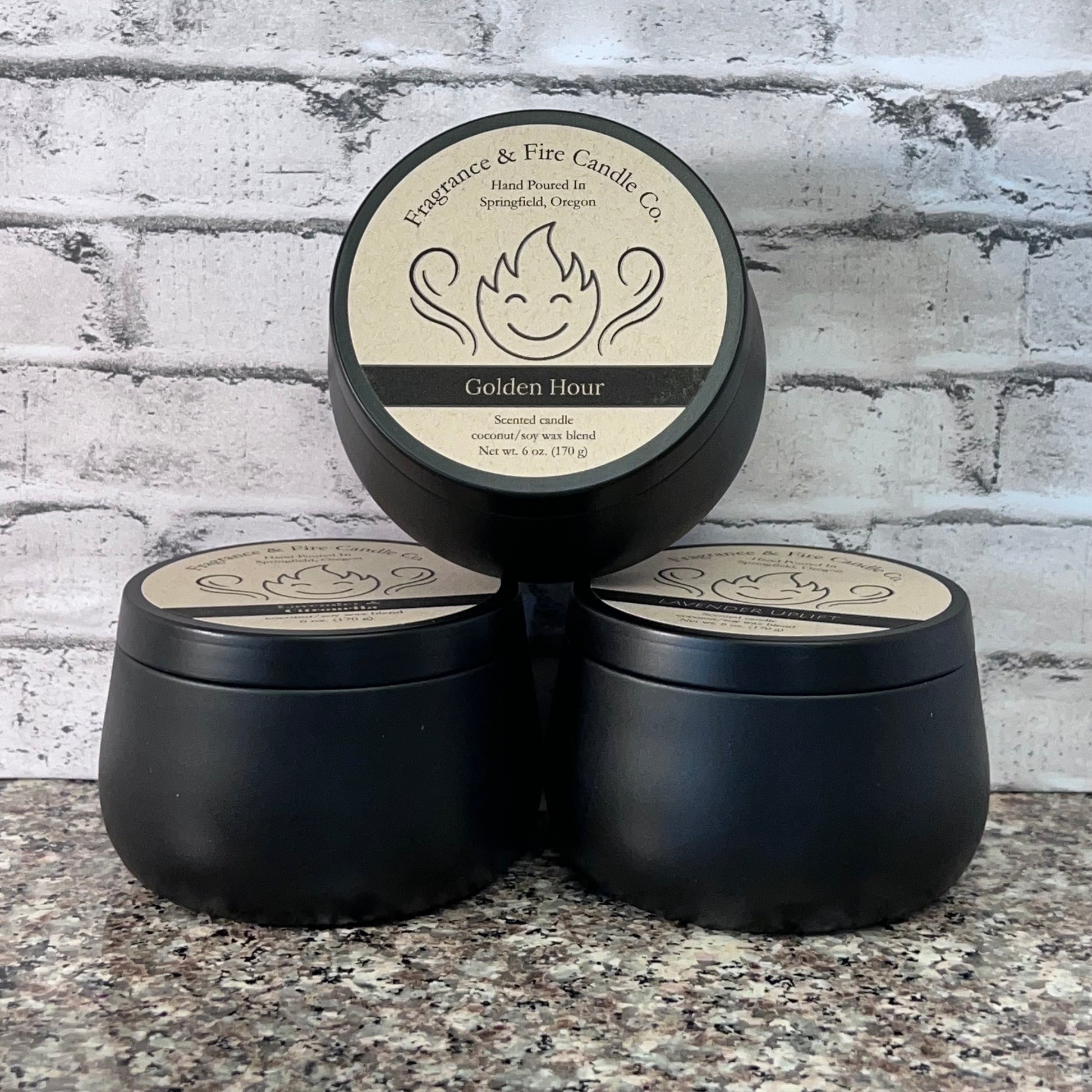 3 Fragrance & Fire candles stacked with 2 on the bottom 1 on top. Top candles is tilted forward so the label shows. Brick wall background.