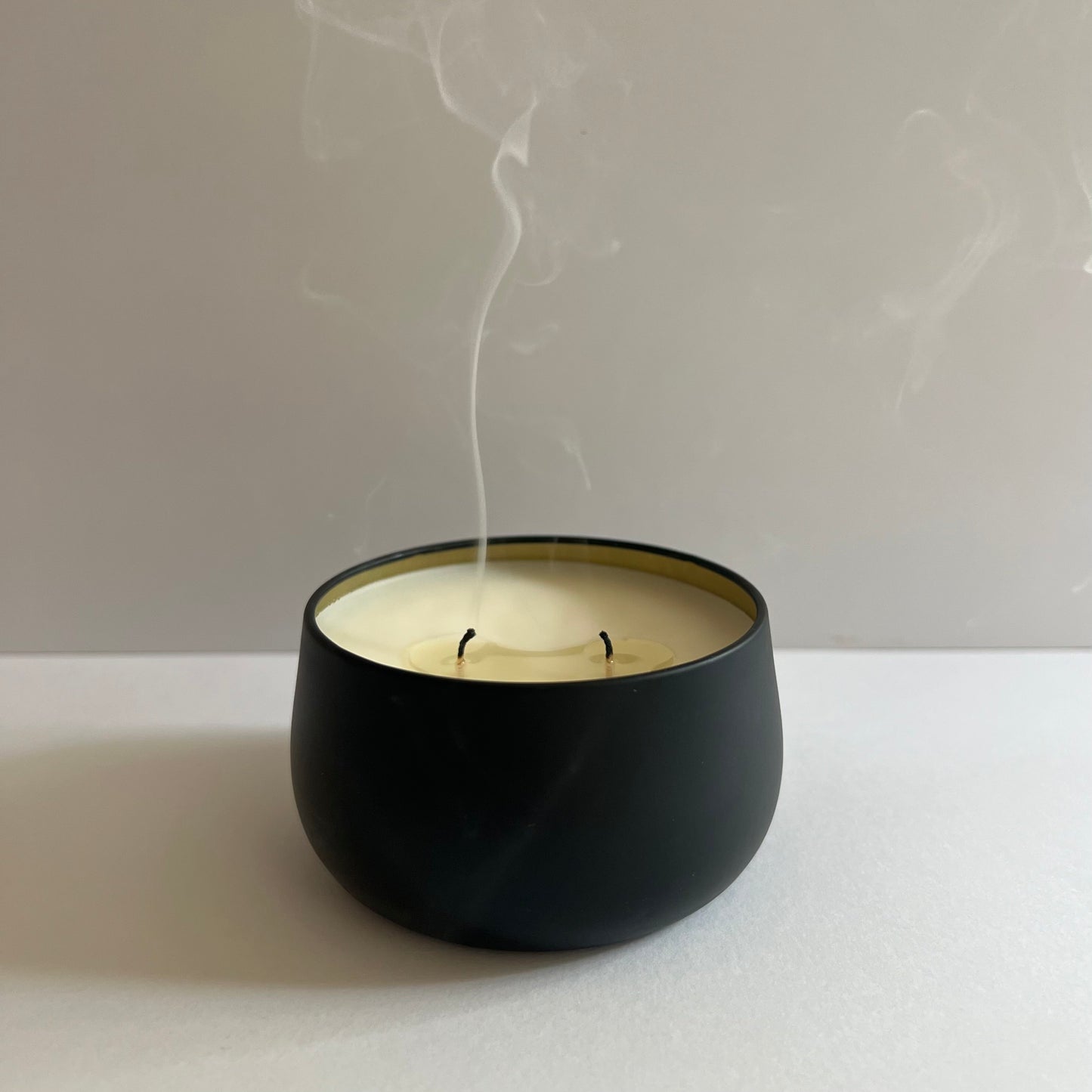 Black container candle, gold lining, 2 wicks just blown out. White smoke rising from wicks. White background.