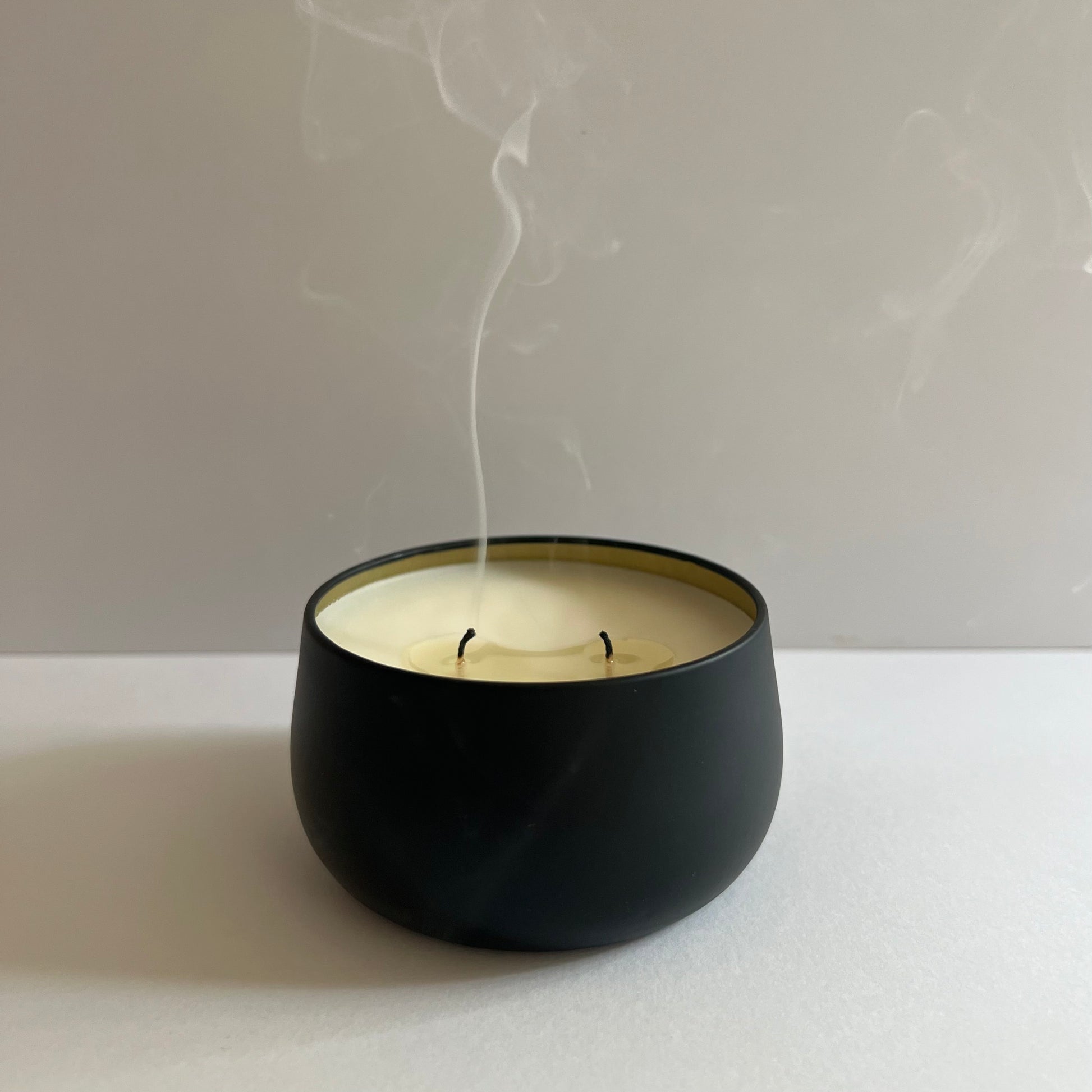 Black container candle, gold lining, 2 wicks just blown out. White smoke rising from wicks. White background.