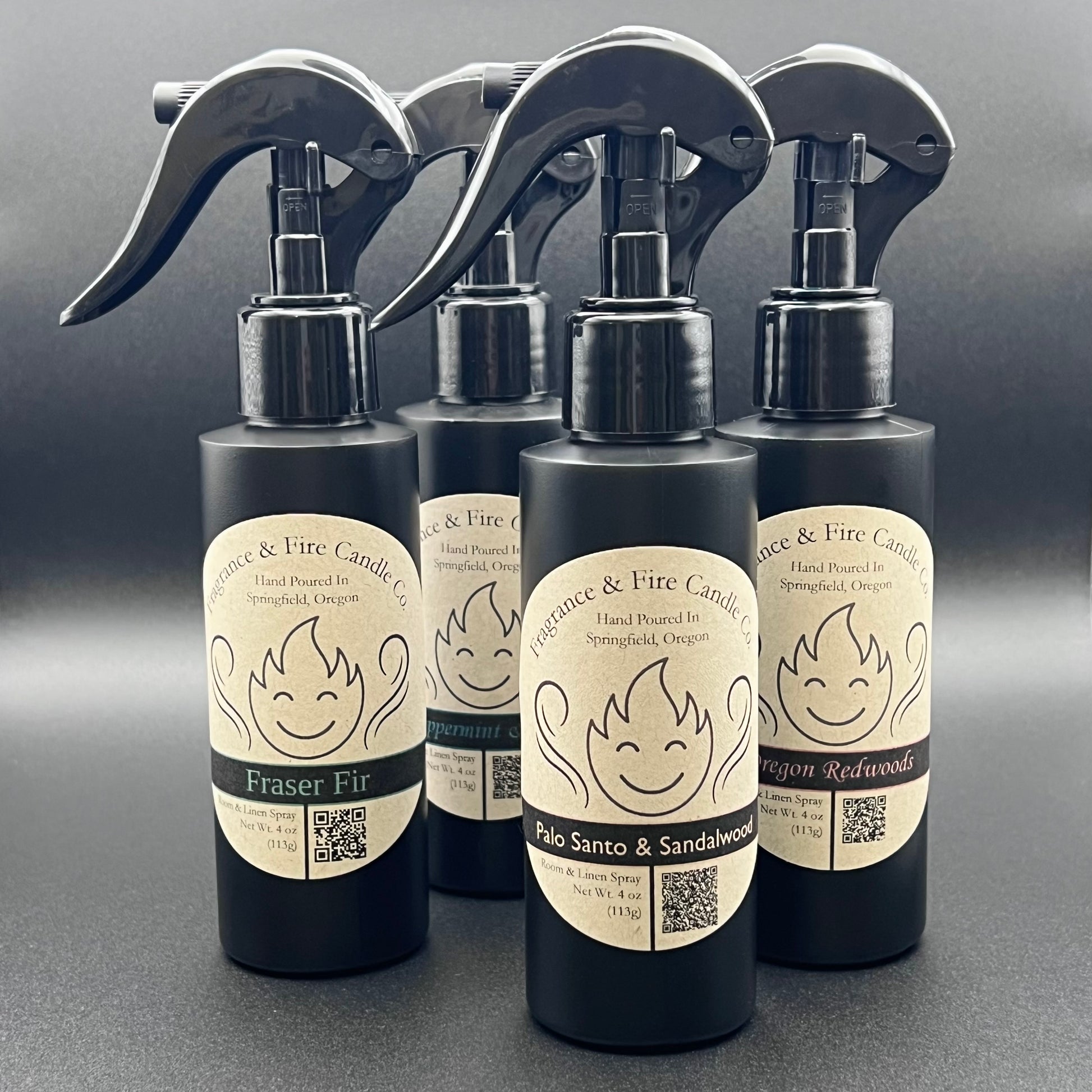 Five black spray bottles from Fragrance & Fire Candle Co., Springfield, Oregon, arranged in a semi-circle against a light black background. Each bottle has a brown label with a stylized flame logo, the text "Fragrance & Fire Candle Co. Hand Poured In Springfield, Oregon," and scent names: "Fraser Fir," "Peppermint & Pine," "Palo Santo & Sandalwood," and "Oregon Redwoods." The labels also include a QR code and the text "Room & Linen Spray Net Wt. 4 oz (113g)." The spray nozzles are black.