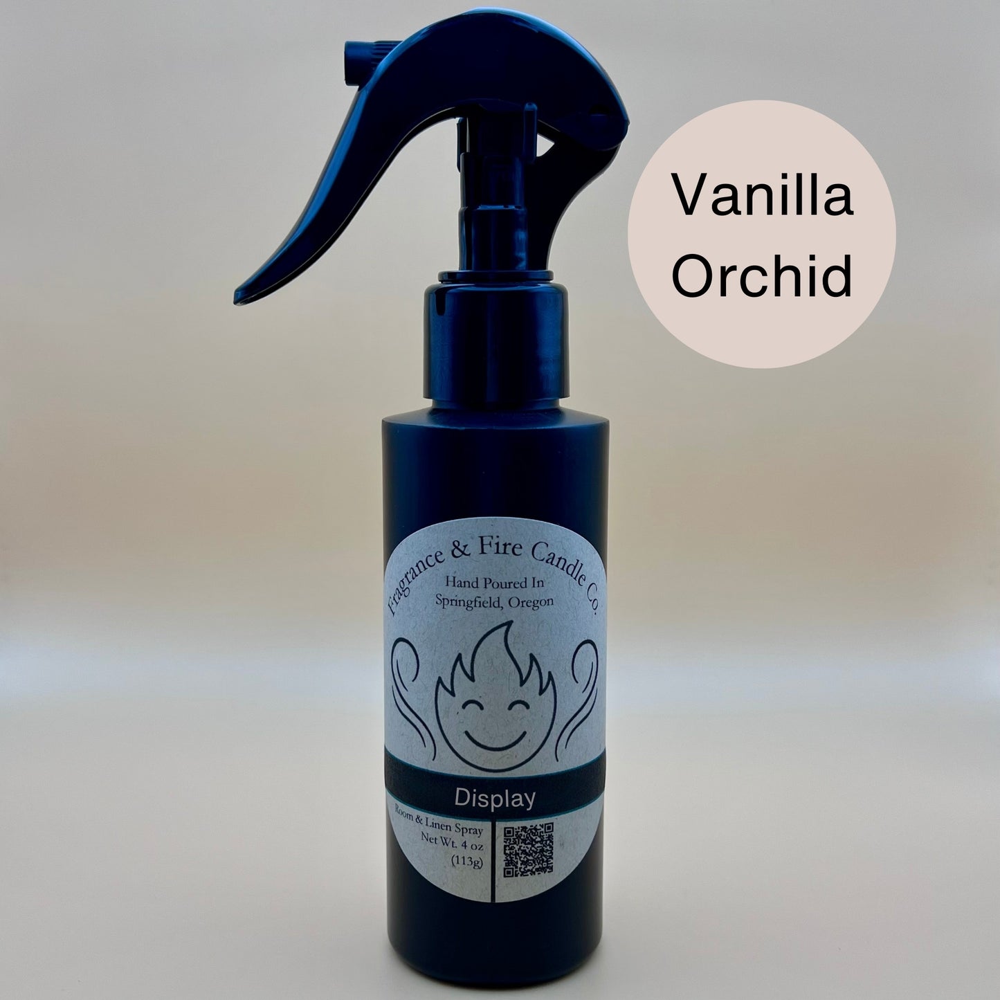 Black spray bottle from Fragrance & Fire Candle Co., Springfield, Oregon, with a black spray nozzle. The bottle has a white label with a stylized flame logo, the text "Fragrance & Fire Candle Co. Hand Poured In Springfield, Oregon," and "Display" scent name. The label also includes a QR code and the text "Room & Linen Spray Net Wt. 4 oz (113g)." A cream circle in the upper right corner reads "Vanilla Orchid."